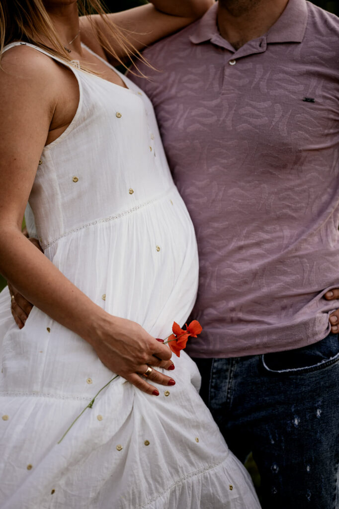 Nahaufnahme. Ein Mann und eine schwangere Frau im weißem Kleid stehen Bauch an Bauch und umarmen sich. Mit der freien Hand hält die Frau eine Mohnblume an ihren Babybauch.